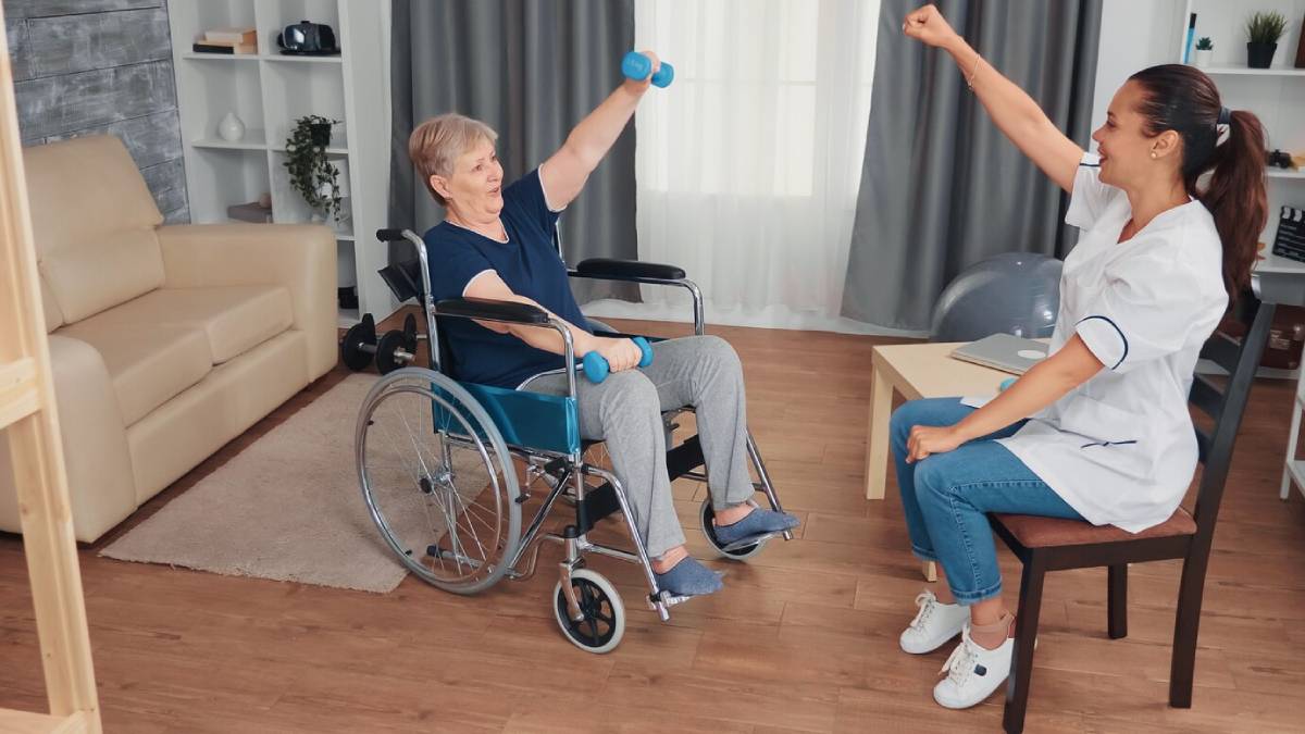 Elderly woman in a wheelchair exercises with dumbbells, cheered on by a smiling nurse in a home setting.