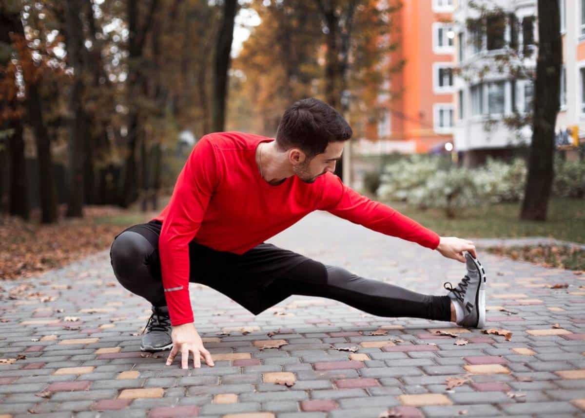 Young man stretching outdoors in a park wearing red sportswear.