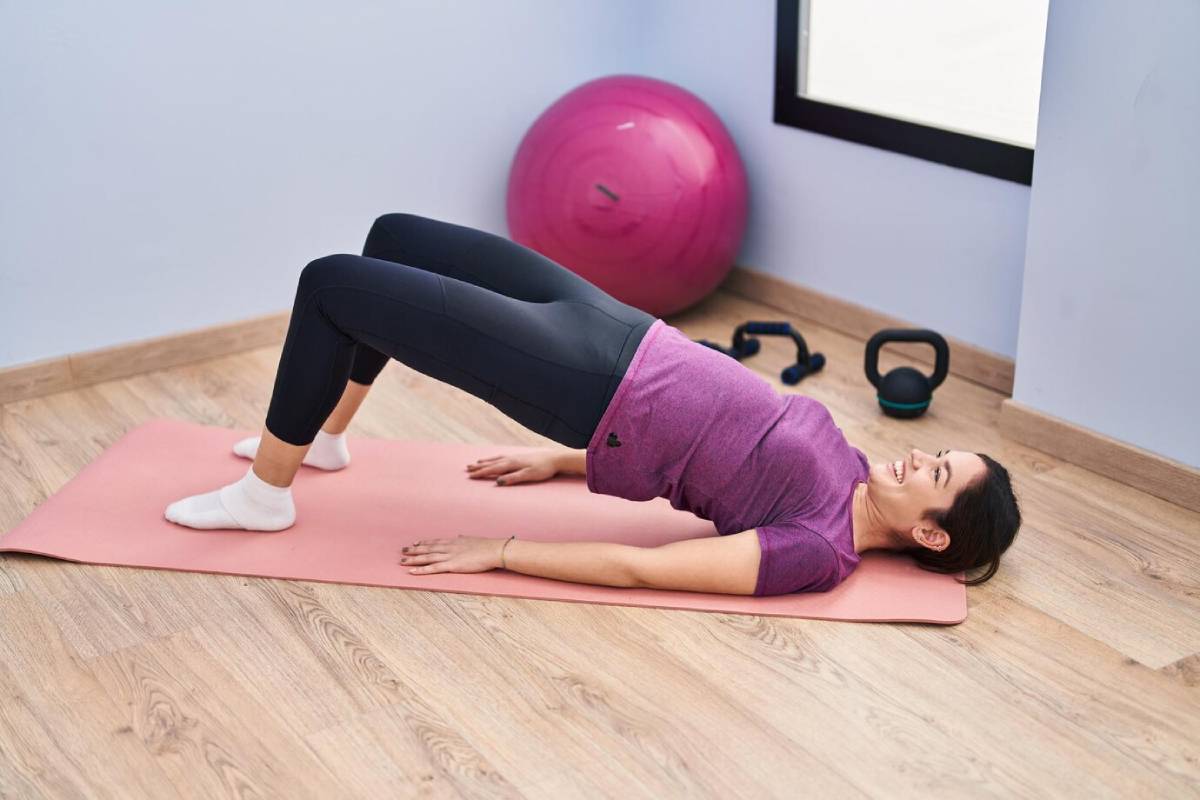 Woman exercising on a mat in a bridge pose, in a home gym setting with fitness equipment in the background.