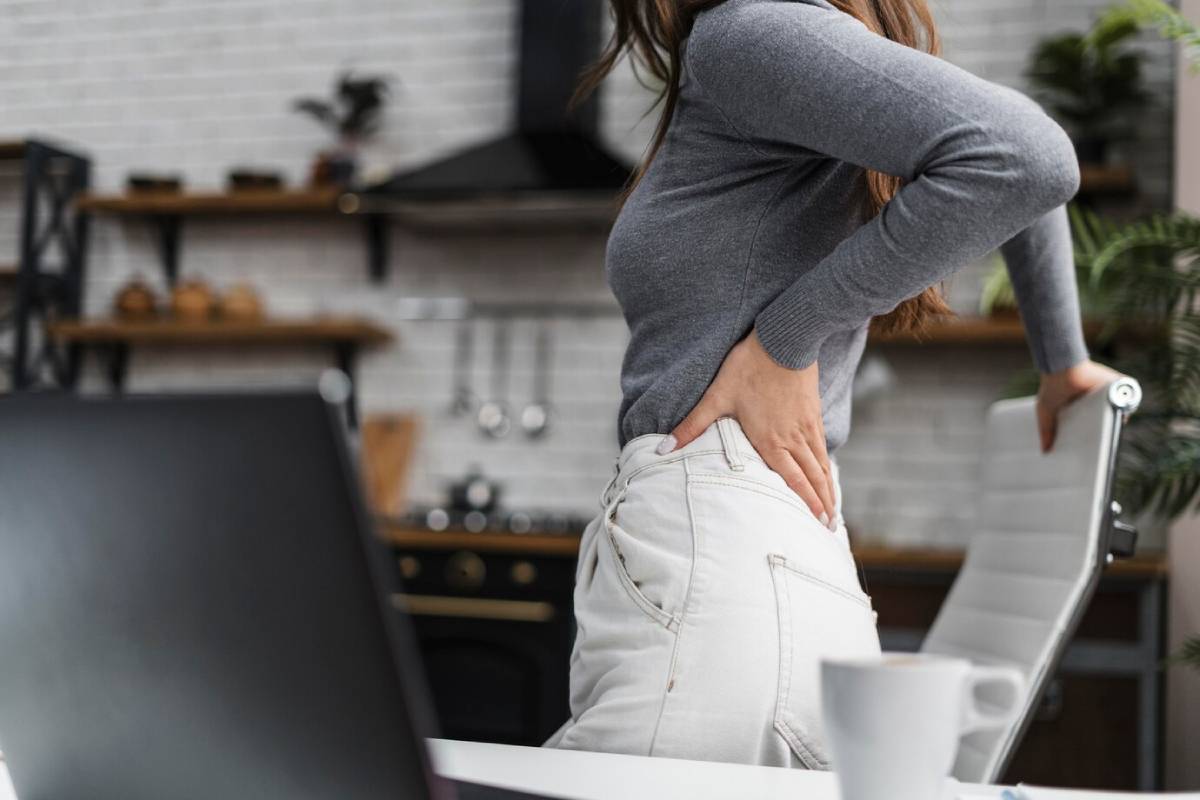 Woman in a grey top, standing by a desk, holding her lower back in a kitchen setting.