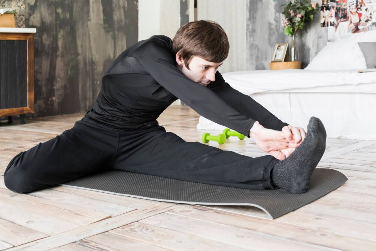 Man in black sports attire stretching on a yoga mat indoors, reaching for his foot.