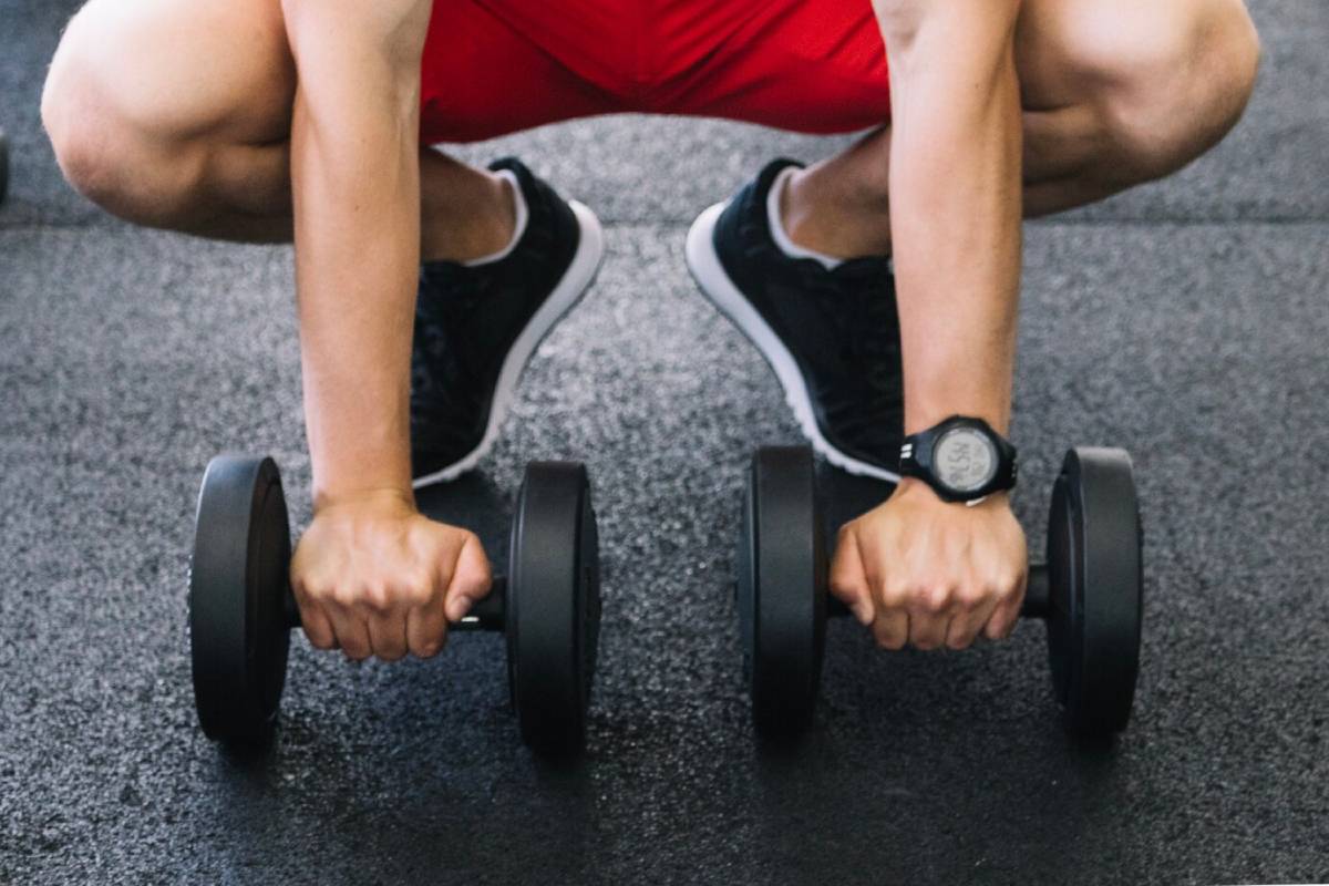 Person in a squat position holding dumbbells, wearing a sports watch and trainers.
