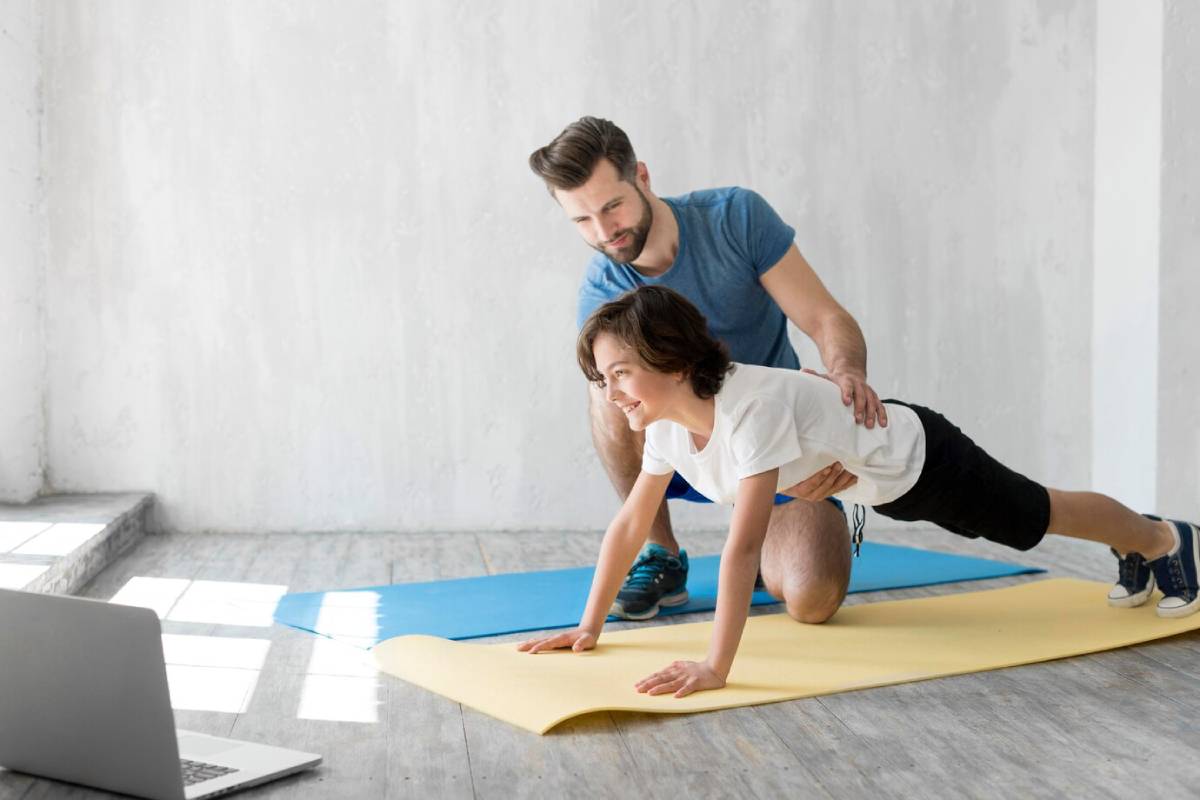 A man assists a young boy with push-ups in a bright room with exercise mats and a laptop nearby.