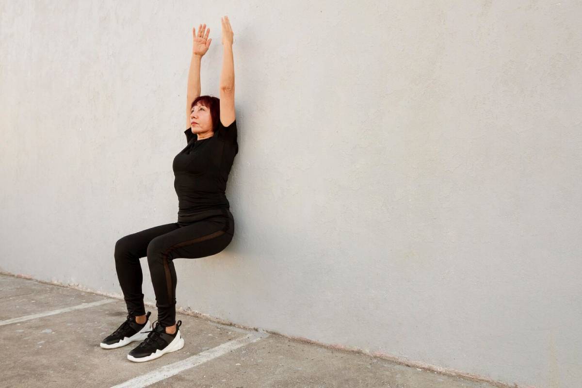 Woman in black exercise outfit doing a wall sit exercise against a plain, light-coloured background.