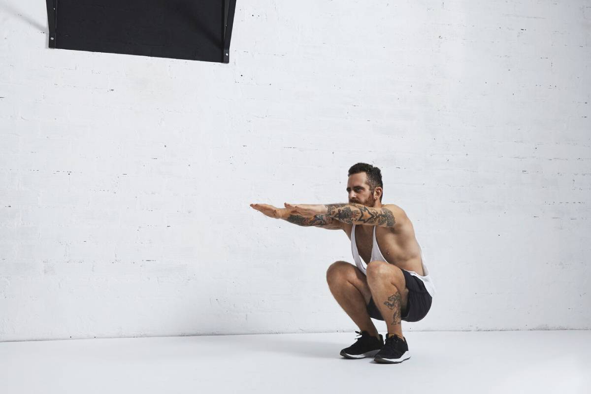 A man with tattoos performs a squat exercise in a gym, extending his arms forward.