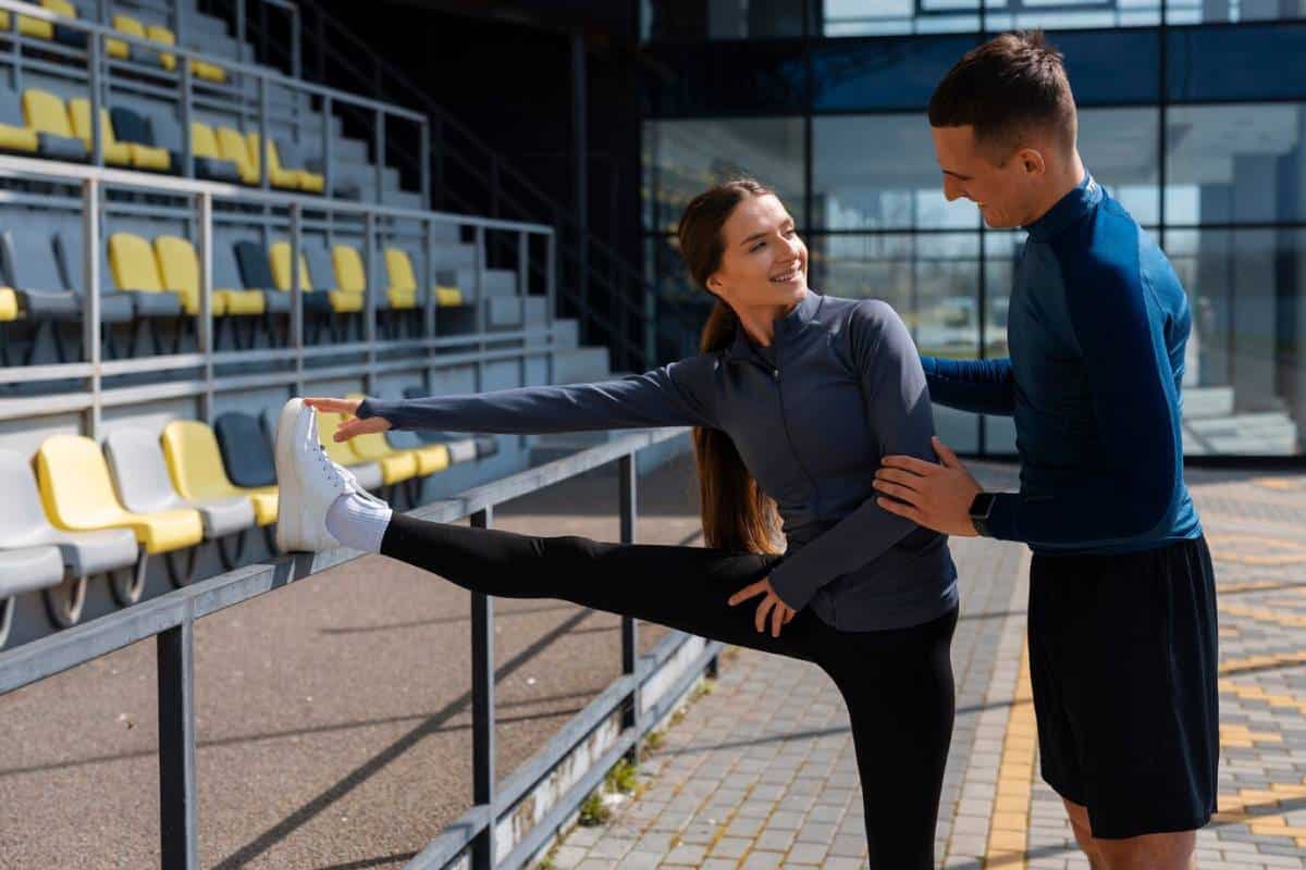A man assists a woman with a leg stretch in a sunny stadium with empty seats.