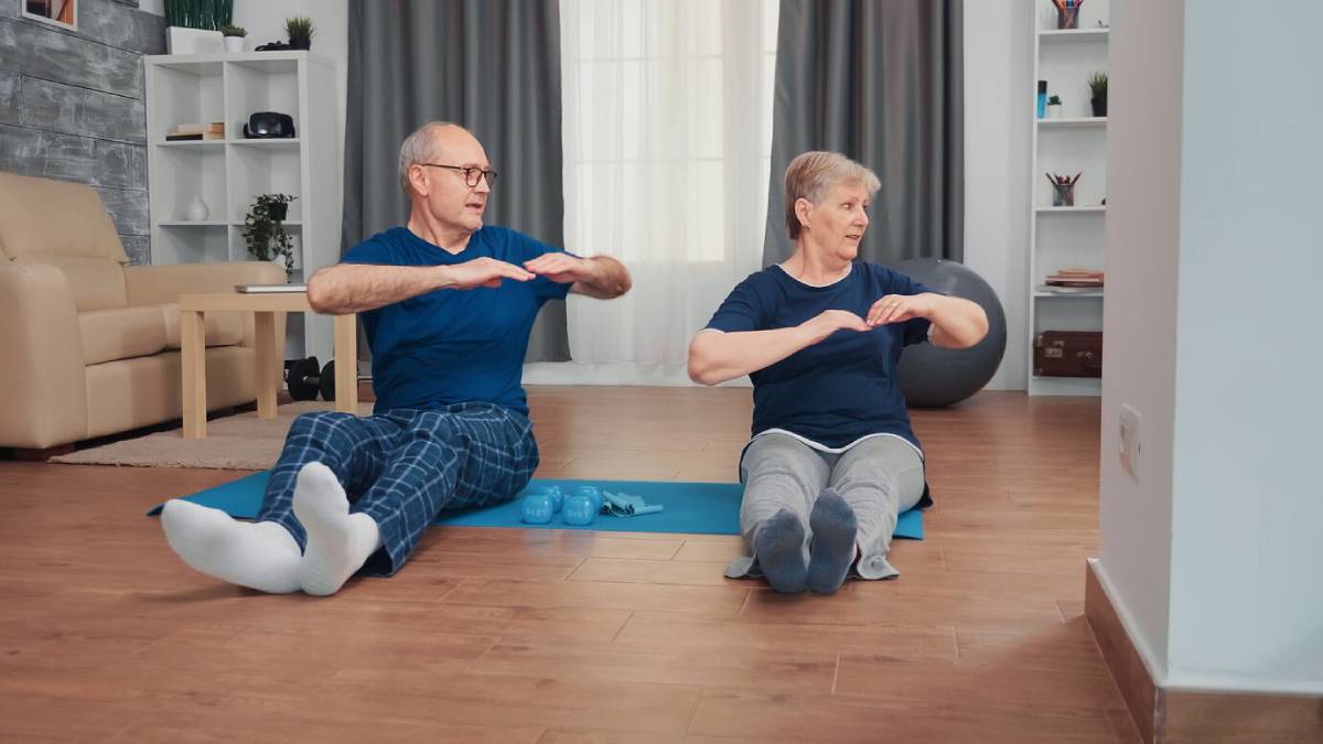 Elderly couple exercises together on mats in a bright living room.