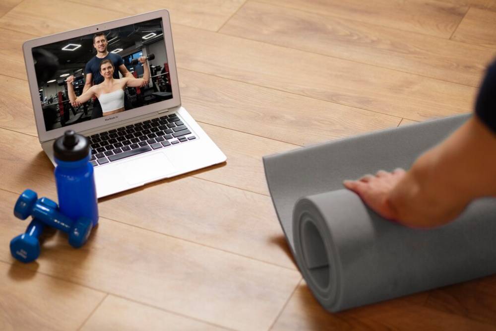  A person unrolls a yoga mat for a virtual workout with instructors displayed on a laptop, alongside a blue water bottle and dumbbells.