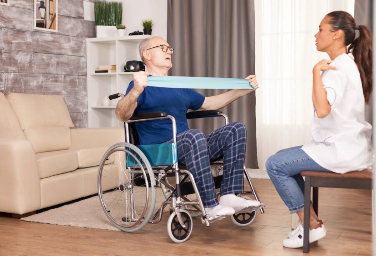 Elderly man in a wheelchair doing physiotherapy exercises with a therapist in a cosy living room.
