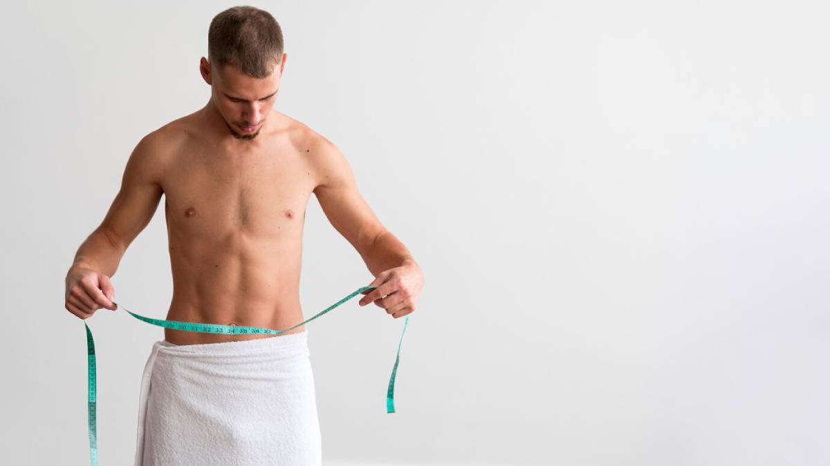 Young man in a towel measuring his waist with a tape measure, looking down at his abdomen.