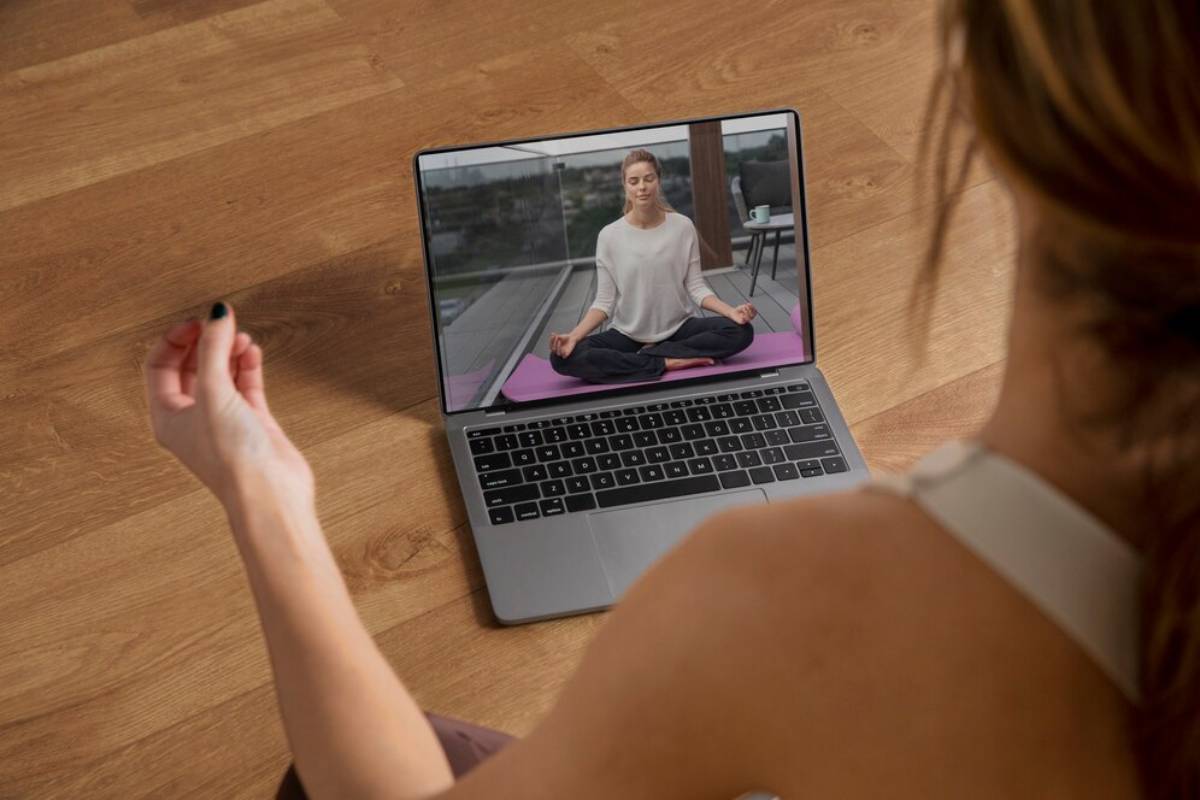 Woman meditating on a yoga mat in an online class viewed on a laptop by another person practicing along.