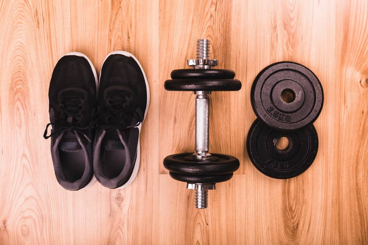 A pair of black running shoes beside a dumbbell and weight plates on a wooden floor.