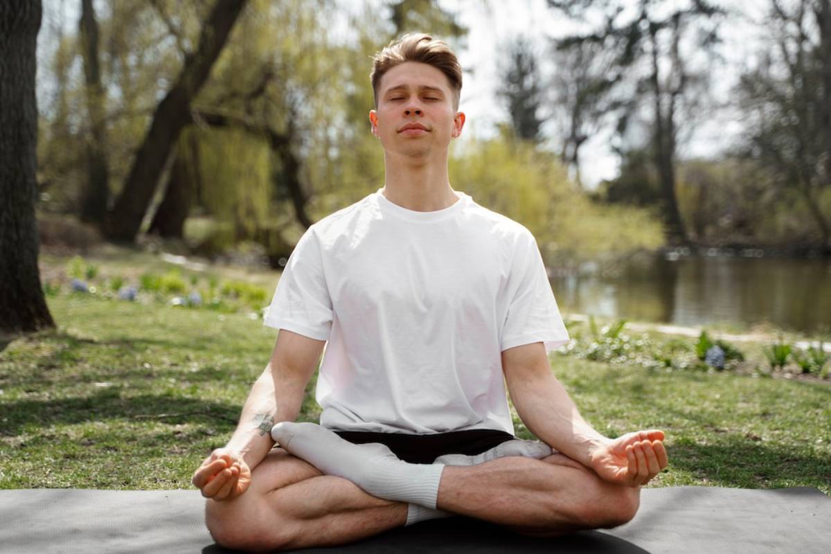 Young man meditating in a cross-legged pose outdoors by a pond, wearing a white shirt and grey shorts.