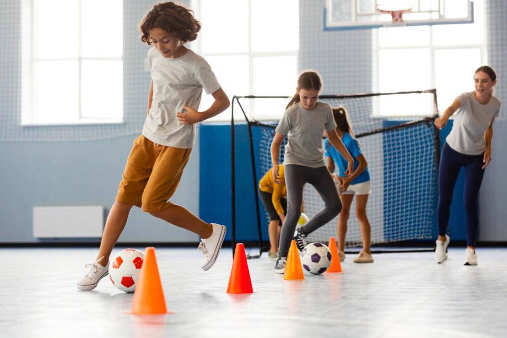  Young boy dribbling a football past orange cones in a sports hall, with onlooking coaches and players.