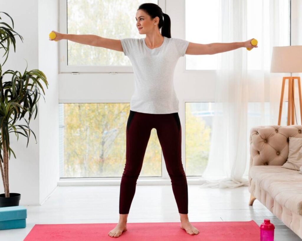 Woman exercising indoors with dumbbells, standing on a yoga mat in a bright room with large windows.