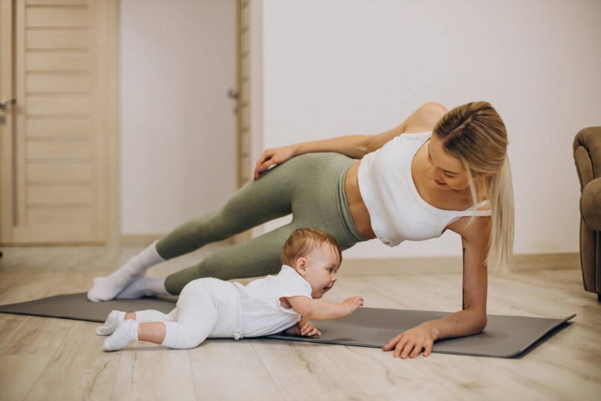 A woman exercises in a side plank position on a yoga mat next to a baby indoors.