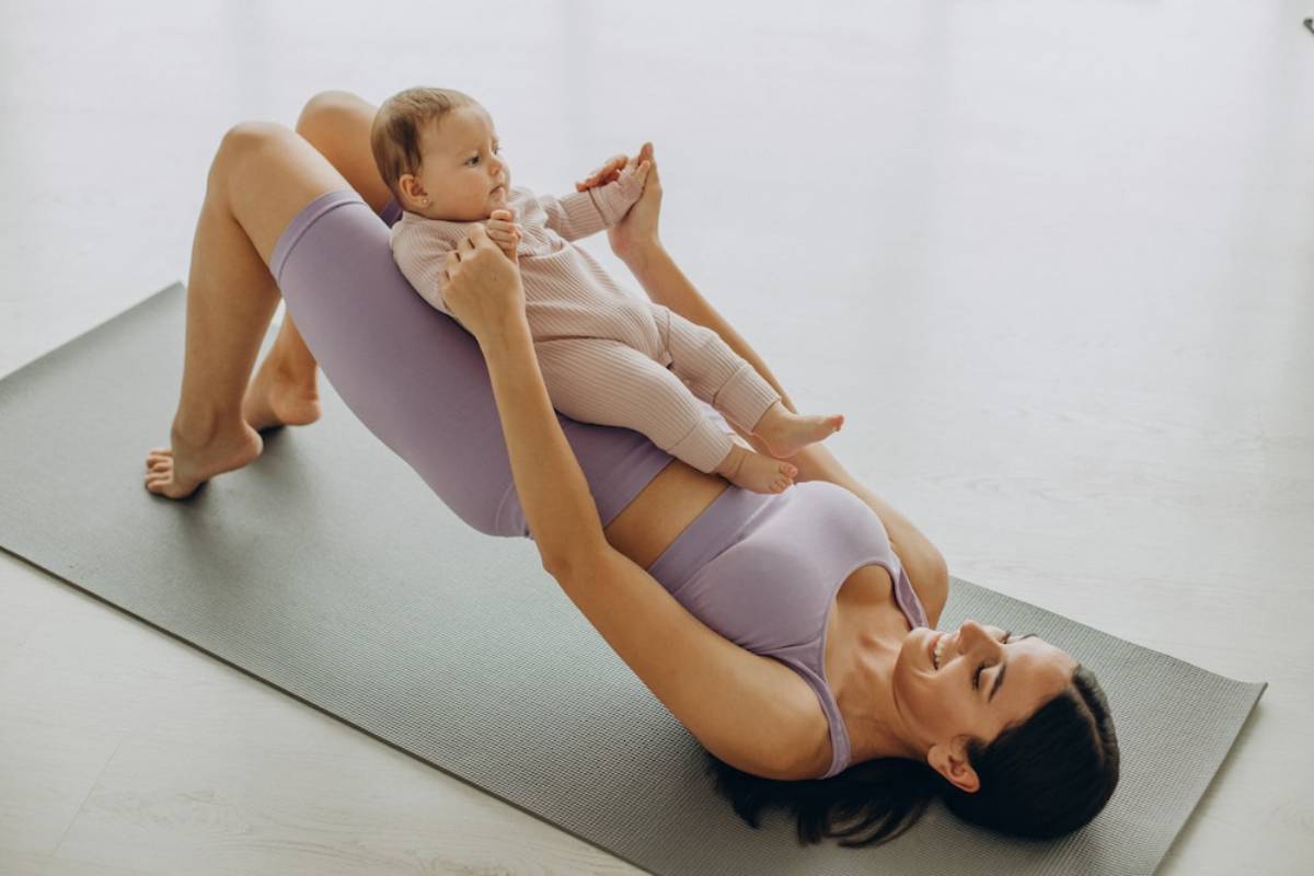 Mother in purple workout attire doing a leg raise exercise on a mat while holding her baby, both smiling.