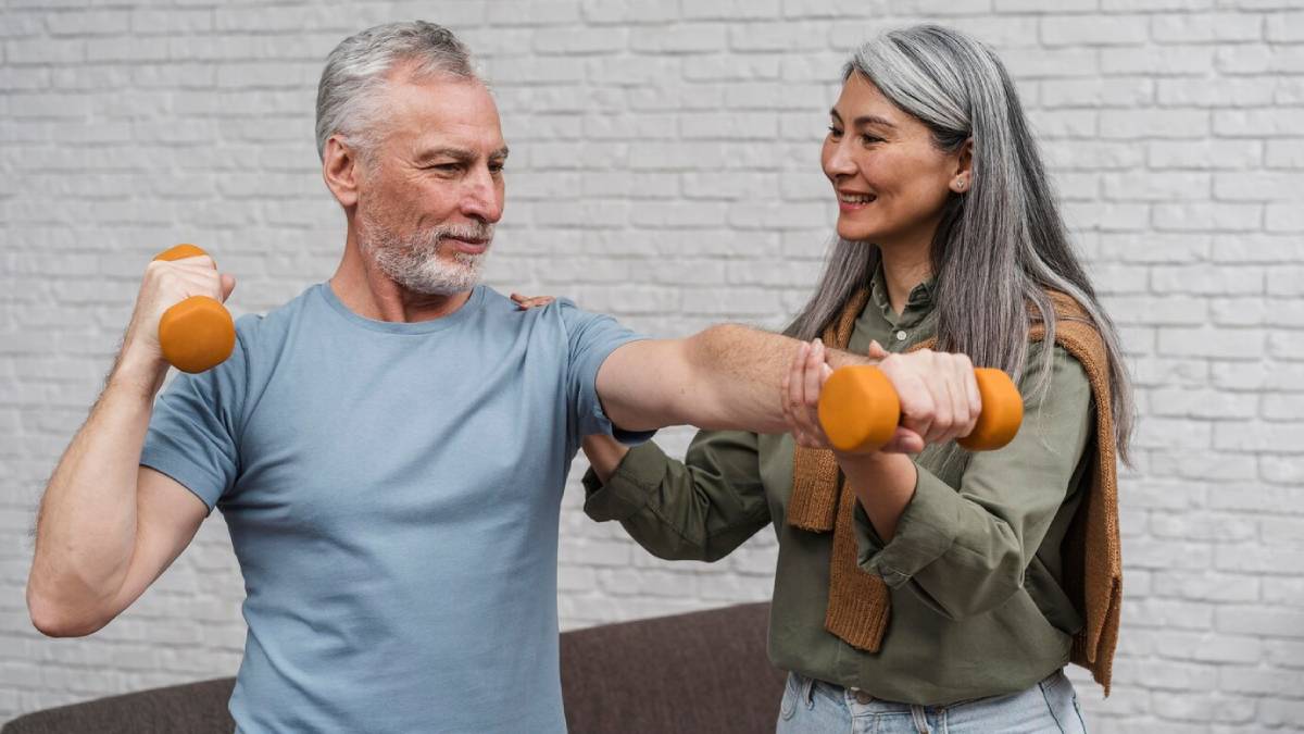 A senior man and woman exercising with dumbbells in a bright room.
