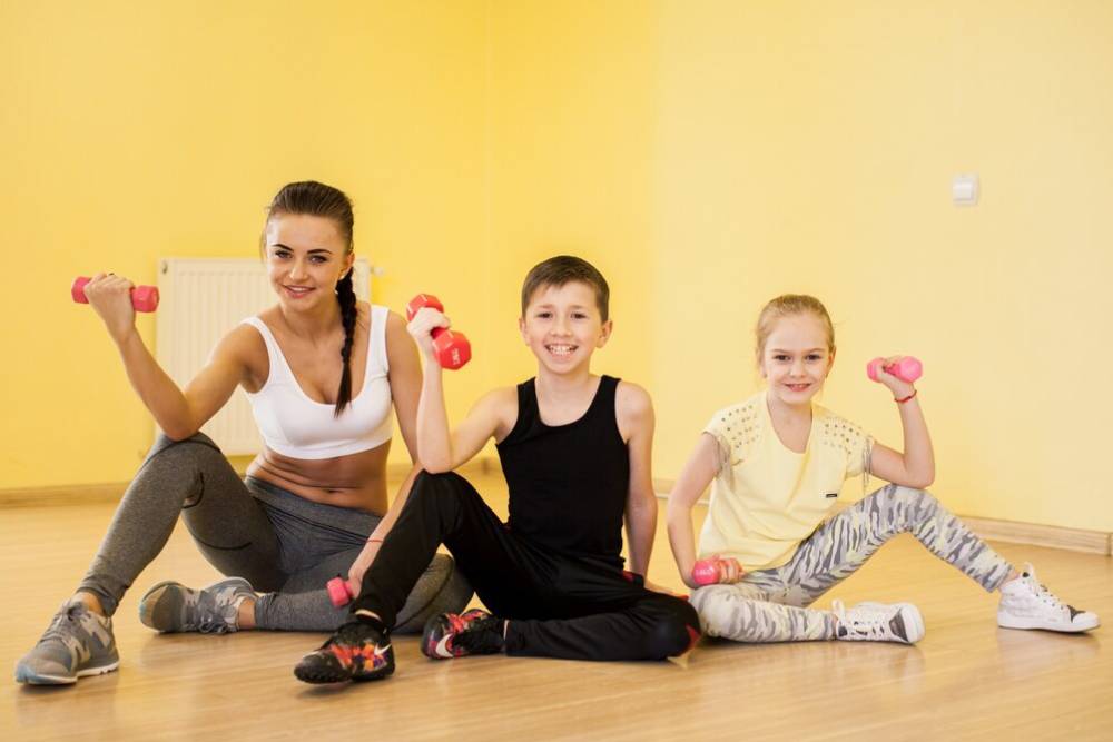  Two children and a woman exercising with dumbbells in a brightly lit room.