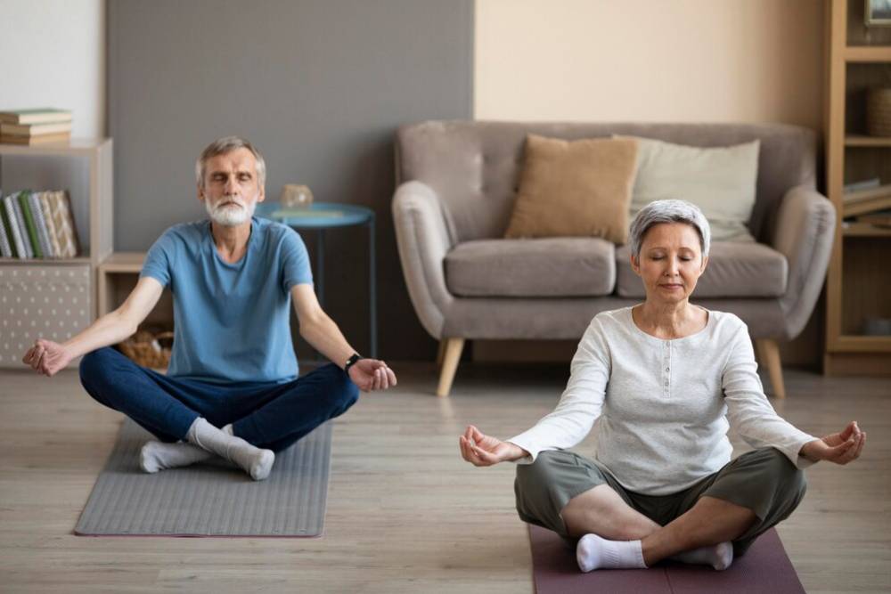 An elderly couple meditates on floor mats in a serene living room.
