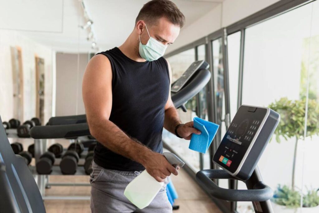 Man in a gym wearing a mask, cleaning an exercise machine with a spray bottle and cloth.