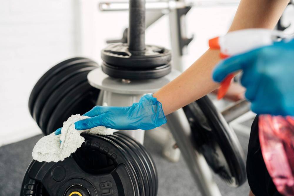  Person in gloves cleaning gym weights with a cloth and spray bottle.