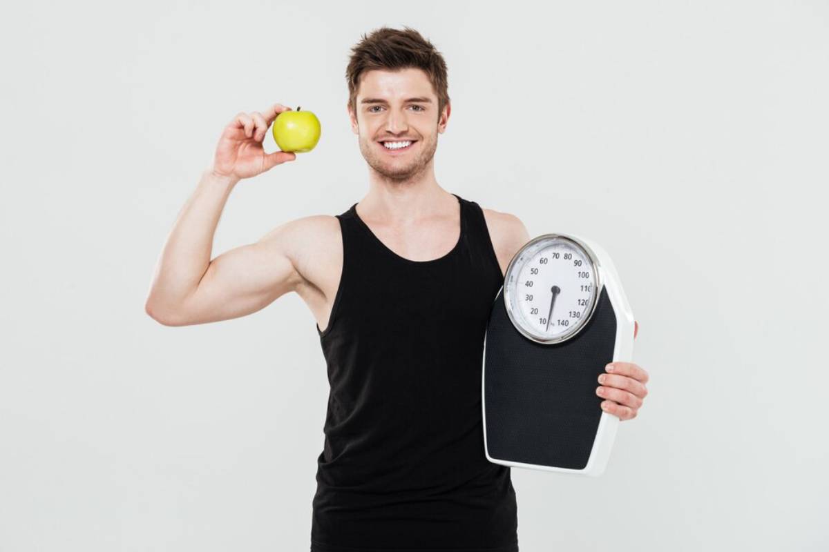 Young man smiling, holding a green apple and a weighing scale, promoting healthy living.