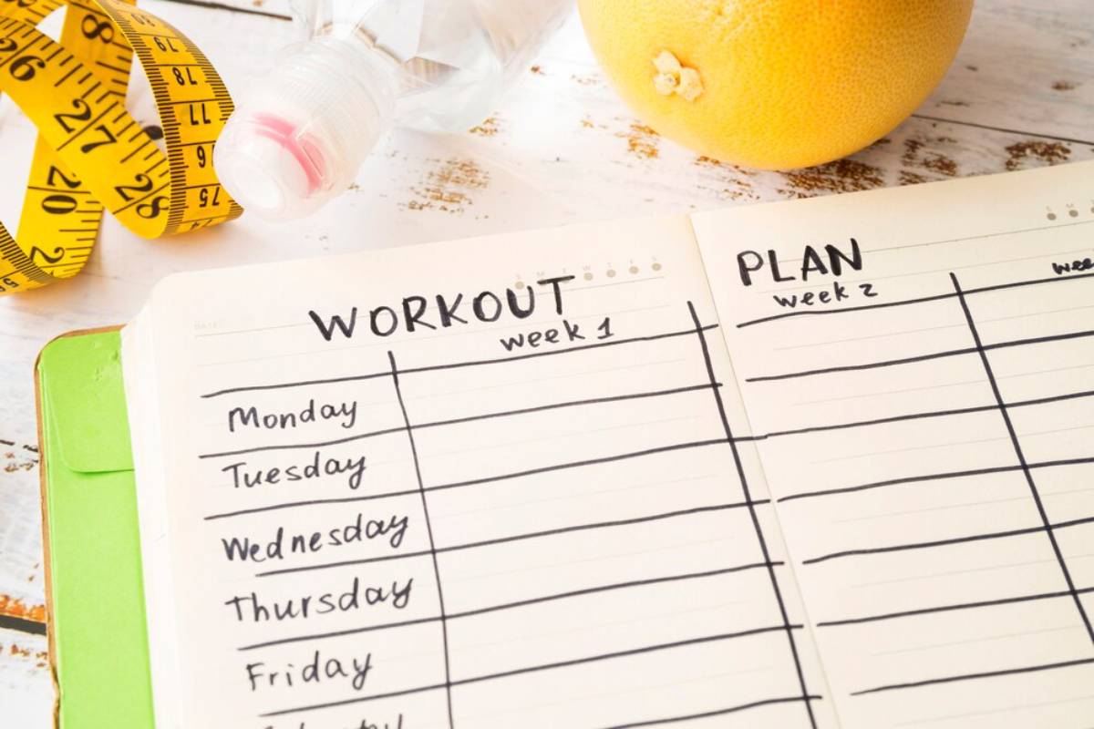 Workout and plan notebook with measuring tape, water bottle, and an orange on a desk.