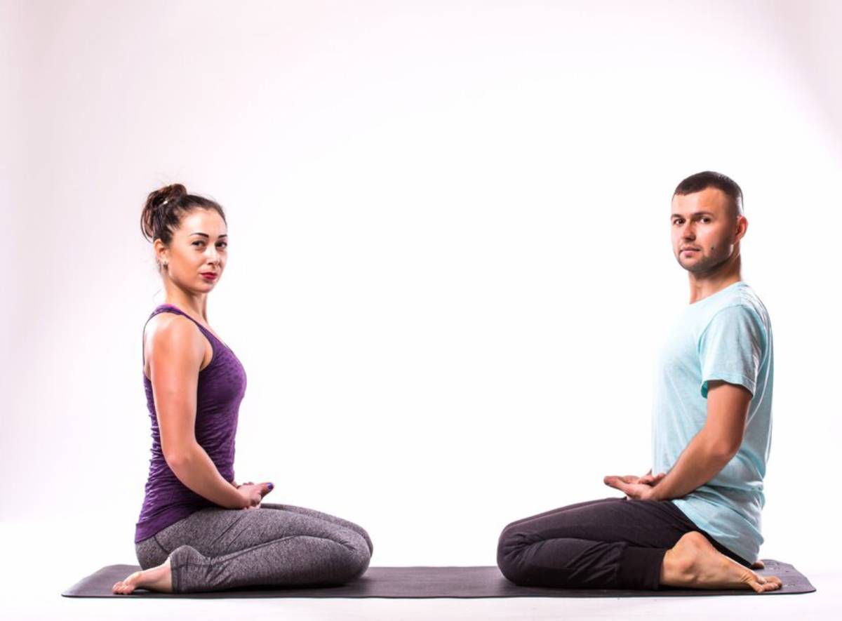 A man and a woman practicing yoga, sitting cross-legged and facing opposite directions against a white background.