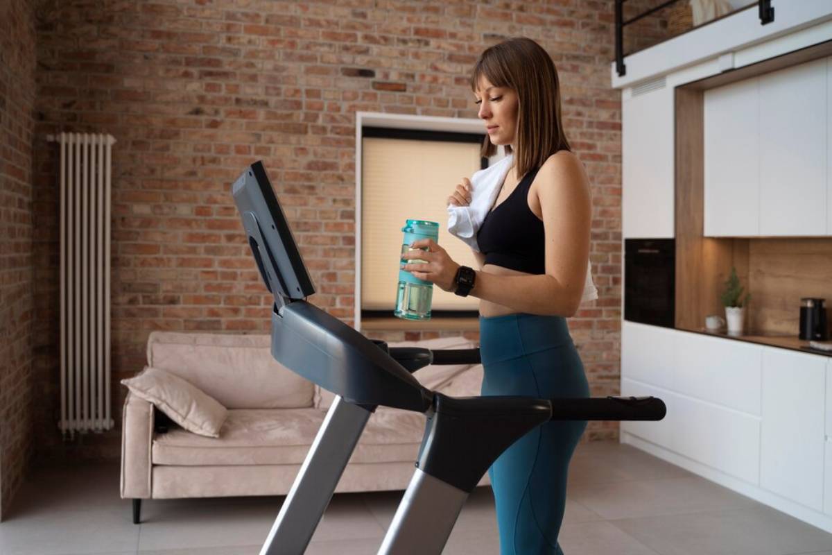 Woman drinking water and resting on a treadmill in a modern home with brick walls.
