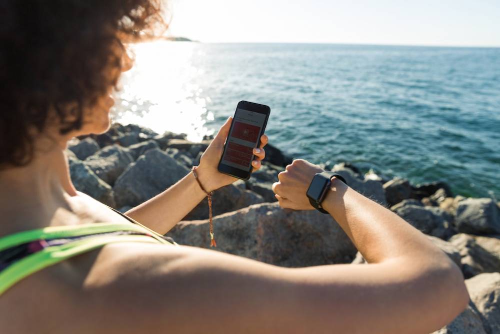 Person checking smartphone on rocky beach, wearing smartwatch, with sunlit sea in background.