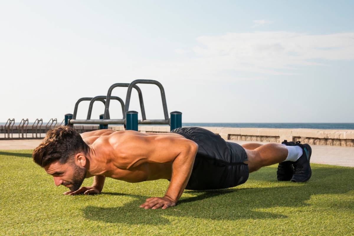 Man performs a push-up outdoor on grass near the seafront, under clear skies.