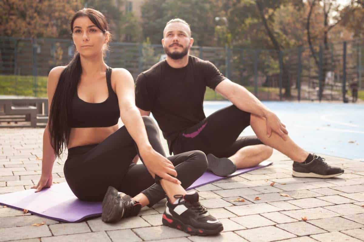 A man and a woman in athletic wear sitting on yoga mats in a park.